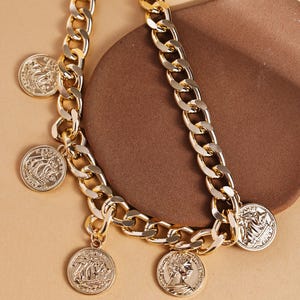 May include: A gold-colored chain necklace with multiple coin pendants. The necklace features a chunky chain design with several circular pendants. Each pendant has a detailed embossed design. The necklace is displayed on a brown surface.