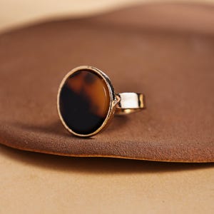 Op de afbeelding: Een goudkleurige ring met een grote, ronde steen met schildpadpatroon. De ring heeft een eenvoudige band en een bezel setting. De steen toont tinten bruin en zwart.