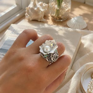 Könnte beinhalten: Ein silberner Ring mit einem Muscheldesign. Der Ring hat eine weiße Muschel mit einer Perle und einem Sternakzent. Das Band hat ein strukturiertes Muschelmuster und kleine, schillernde Edelsteine. Der Ring befindet sich an einem Finger.