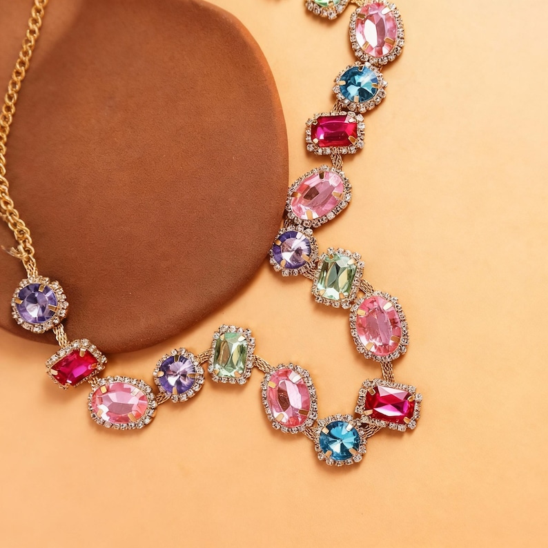 May include: A gold-toned necklace featuring a series of colorful, gem-like stones in various shapes. The stones are in shades of pink, blue, green, and purple, and are set with small, clear crystals. The necklace is displayed on a brown surface.