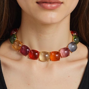 May include: A colorful necklace featuring large, square-cut gemstones in a variety of colors, including red, orange, yellow, pink, green, and purple. The gemstones are set in gold-colored metal. The necklace is a statement piece.