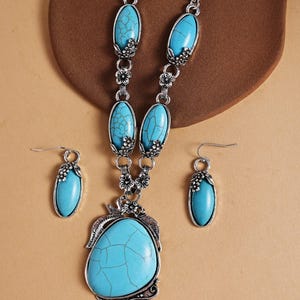 May include: A silver-toned jewelry set featuring a necklace and matching earrings. The necklace has a large, turquoise-colored pendant and smaller turquoise stones. The earrings have a similar turquoise stone design. Floral accents are incorporated throughout the set.