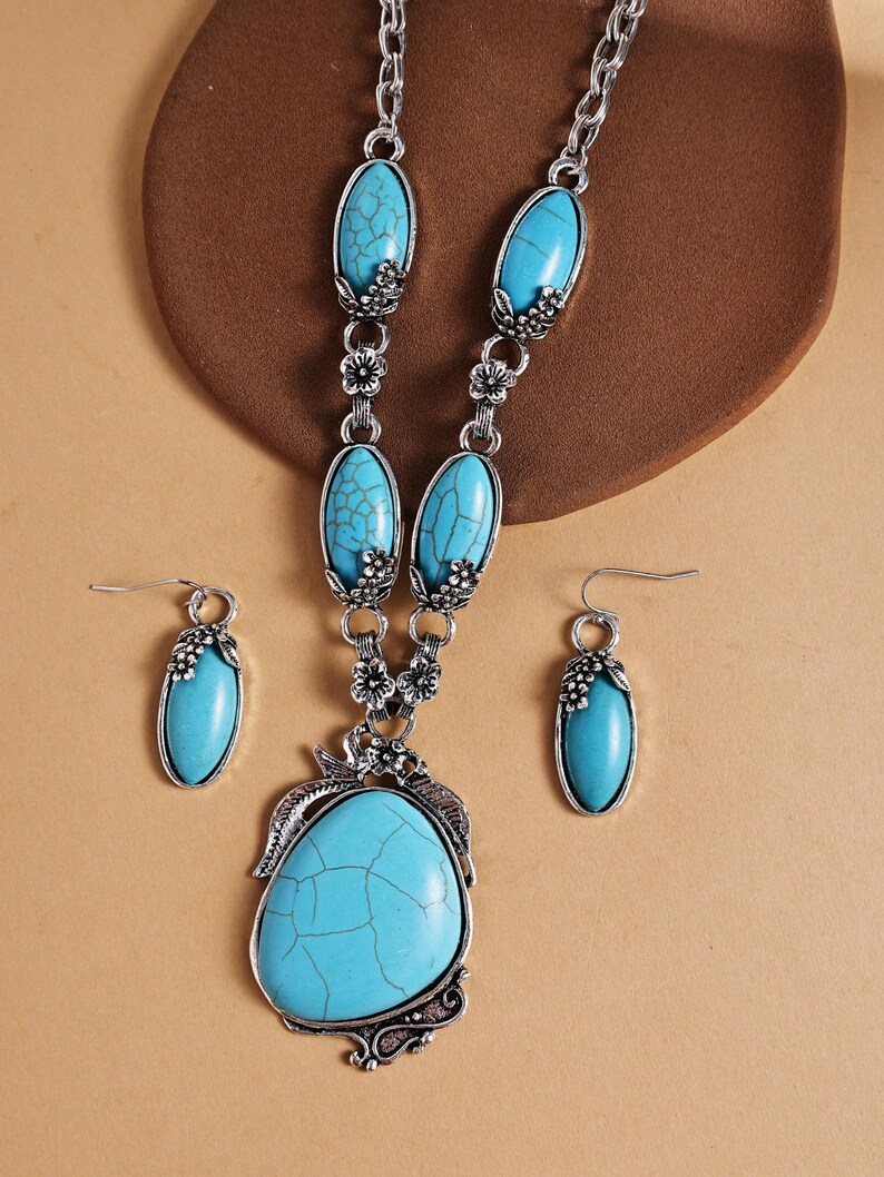 May include: A silver-toned jewelry set featuring a necklace and matching earrings. The necklace has a large, turquoise-colored pendant and smaller turquoise stones. The earrings have a similar turquoise stone design. Floral accents are incorporated throughout the set.
