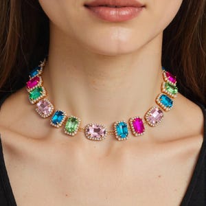 May include: A colorful statement necklace featuring rectangular gemstones in shades of pink, blue, green, and fuchsia. The gems are set in a gold-tone metal with a halo of small, clear crystals. The necklace is a bold and eye-catching piece of jewelry.
