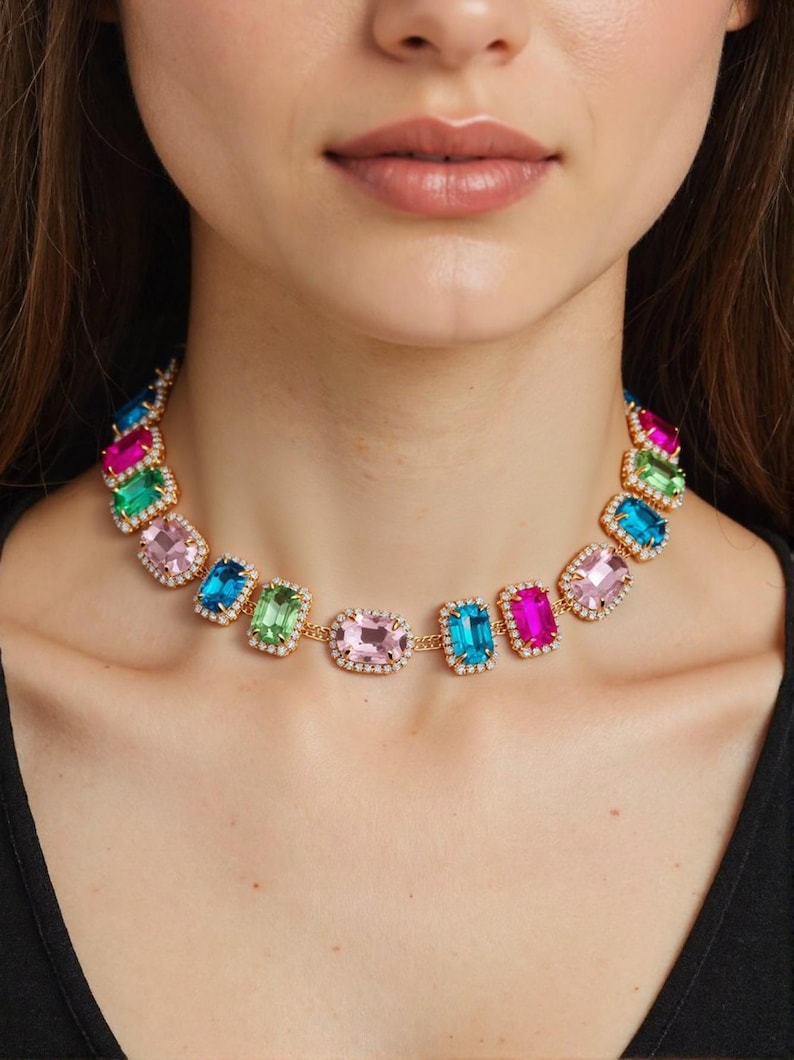 May include: A colorful statement necklace featuring rectangular gemstones in shades of pink, blue, green, and fuchsia. The gems are set in a gold-tone metal with a halo of small, clear crystals. The necklace is a bold and eye-catching piece of jewelry.