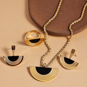 Op de afbeelding: Een goudkleurige sieradenset met een ketting, oorbellen, ring en hanger. Elk stuk heeft een halve cirkel ontwerp met een zwart midden. De ketting heeft een touwketting en de oorbellen zijn oorstekers.