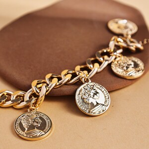 May include: Gold-toned chain bracelet with coin charms. The bracelet features a thick chain design with several circular coin pendants. The coins have a detailed relief of a classical figure. The bracelet is displayed on a brown surface.