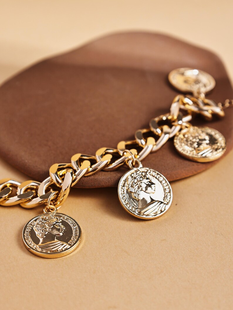 May include: Gold-toned chain bracelet with coin charms. The bracelet features a thick chain design with several circular coin pendants. The coins have a detailed relief of a classical figure. The bracelet is displayed on a brown surface.
