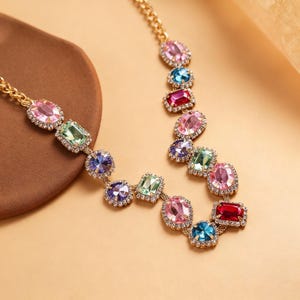 May include: A gold-toned necklace featuring a series of colorful, gem-like stones in various shapes. The stones are in shades of pink, green, blue, purple, and red, set in a gold-colored metal. The necklace is displayed on a neutral background.