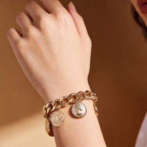 May include: Gold-toned chain bracelet with coin charms. The bracelet features a thick, curb chain design. Several circular coin charms dangle from the chain, each with a detailed relief design. The bracelet is worn on a wrist against a neutral background.