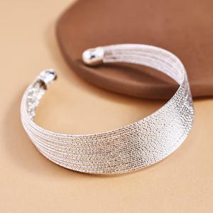 May include: A silver cuff bracelet with a textured finish. The bracelet has a curved design and is open at one end. The cuff is displayed on a light brown surface.