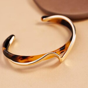 Peut inclure: Un bracelet manchette dor&eacute; avec un motif &eacute;caille de tortue. Le bracelet a un design incurv&eacute; et ouvert avec une finition lisse et polie. Le motif &eacute;caille de tortue pr&eacute;sente des nuances de marron et de noir.