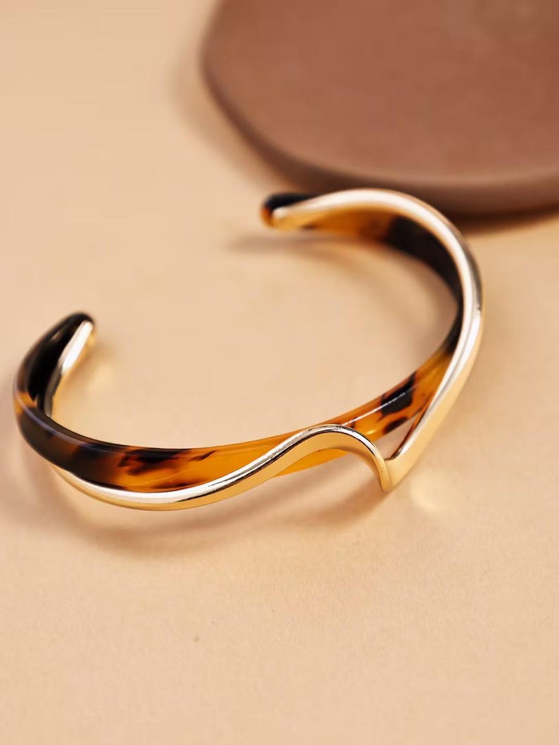 Peut inclure: Un bracelet manchette dor&eacute; avec un motif &eacute;caille de tortue. Le bracelet a un design incurv&eacute; et ouvert avec une finition lisse et polie. Le motif &eacute;caille de tortue pr&eacute;sente des nuances de marron et de noir.