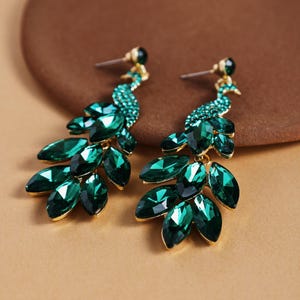 Könnte beinhalten: Ein Paar smaragdgrüne Ohrhänger. Jeder Ohrring hat ein Pfauendesign mit goldfarbenem Metallfinish. Die Ohrringe sind mit grünen Edelsteinen in einem blattartigen Muster verziert, was ein glamouröses Accessoire ergibt.