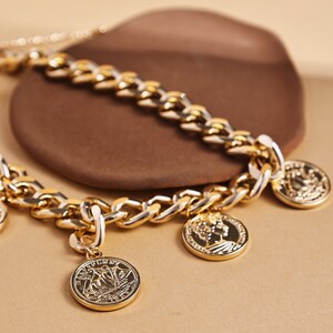 May include: A gold-toned chain necklace with multiple coin pendants. The necklace features a chunky chain design with detailed coin charms. The coins have intricate embossed designs. The necklace is displayed on a neutral background.