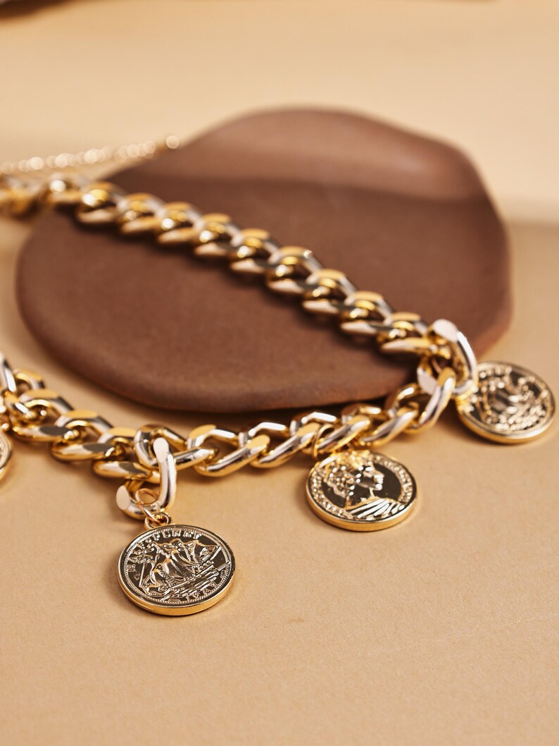 May include: A gold-toned chain necklace with multiple coin pendants. The necklace features a chunky chain design with detailed coin charms. The coins have intricate embossed designs. The necklace is displayed on a neutral background.
