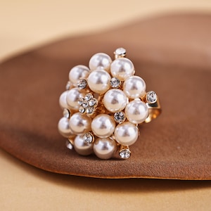May include: A gold ring adorned with a cluster of faux pearls and clear crystals. The white pearls are spherical, forming a textured, floral pattern. The ring is set on a brown surface. The ring's design is reminiscent of a flower.