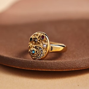 Könnte beinhalten: Ein goldfarbener Ring mit ovaler Oberfläche, der Himmelsmotive und farbige Edelsteine aufweist. Die Ringoberfläche zeigt einen Stern, einen Mond und ein Auge mit einer Vielzahl kleiner, farbiger Steine. Der Ring liegt auf einer braunen Oberfläche.