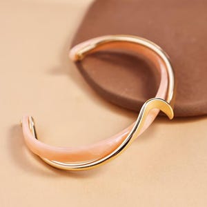 Könnte beinhalten: Ein goldfarbenes Armband mit geschwungenem Design. Das Armband hat eine hellrosa Emaille-Beschichtung und eine polierte Goldoberfläche. Der Schmuck wird vor einem neutralen Hintergrund präsentiert, der seine elegante Form hervorhebt.