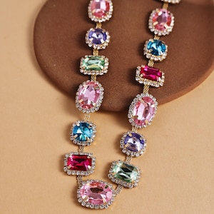 May include: A gold-toned necklace featuring a series of colorful, gem-like stones in various shapes. Each stone is framed with small, clear crystals. The necklace displays a range of colors, including pink, blue, green, and purple, creating a vibrant and eye-catching accessory.