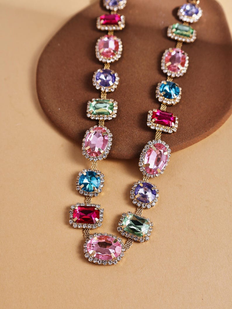 May include: A gold-toned necklace featuring a series of colorful, gem-like stones in various shapes. Each stone is framed with small, clear crystals. The necklace displays a range of colors, including pink, blue, green, and purple, creating a vibrant and eye-catching accessory.