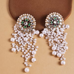 May include: A pair of gold-toned earrings with a floral design, featuring clear and green gemstones, surrounded by small pearls. White pearl strands cascade from the earrings, creating a dangling effect. The earrings are set against a neutral background.