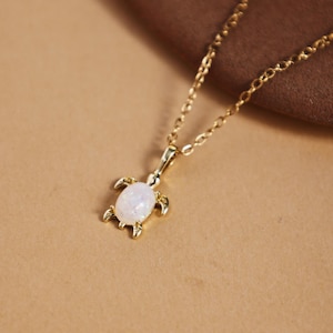 Op de afbeelding: Een goudkleurige ketting met een schildpadhanger met een witte, iriserende steen. De schildpadhanger heeft gedetailleerde poten en een getextureerd schild. De ketting is delicaat.