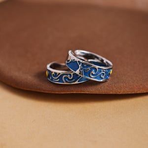 May include: Two silver rings with a blue enamel design featuring a swirling pattern and yellow accents. The rings are set against a brown background, showcasing the intricate details of the jewellery.
