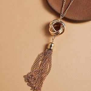 Op de afbeelding: Goudkleurige ketting met een geknoopte hanger en een kettingkwast. De ketting heeft een fijne ketting en een gepolijste afwerking. De kwast is gemaakt van meerdere strengen fijne kettingen, wat een vloeiend effect creëert. De achtergrond is effen neutraal.