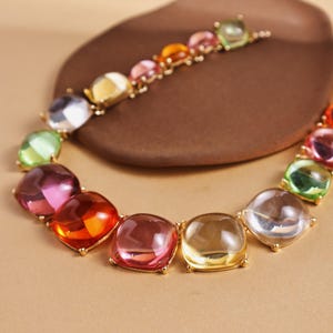 May include: A colorful necklace featuring large, square-cut gemstones in shades of pink, orange, yellow, green, and clear. The gemstones are set in a gold-colored metal and arranged in a circular pattern. The necklace is displayed on a brown, organic-shaped surface.