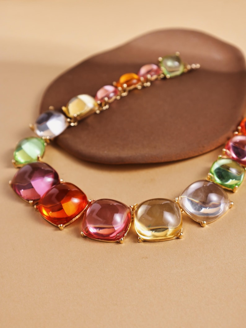 May include: A colorful necklace featuring large, square-cut gemstones in shades of pink, orange, yellow, green, and clear. The gemstones are set in a gold-colored metal and arranged in a circular pattern. The necklace is displayed on a brown, organic-shaped surface.