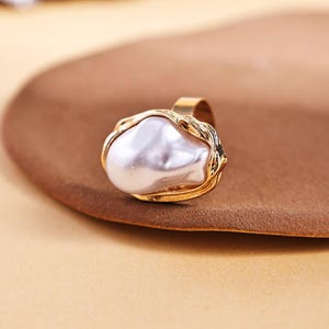 May include: A gold-toned ring featuring a large, irregularly shaped white pearl. The pearl is set within a textured gold band, creating a striking contrast. The ring sits on a brown surface.