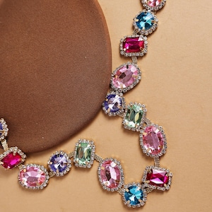 May include: A gold-toned necklace featuring a series of colorful, faceted gemstones in various shapes. The gemstones are set in a delicate, linked chain, with a mix of pink, blue, green, and purple hues. The necklace is displayed on a brown surface.