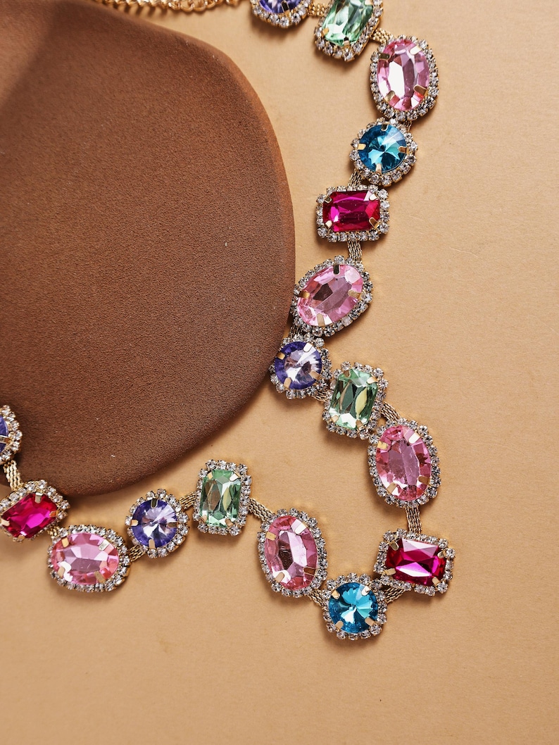 May include: A gold-toned necklace featuring a series of colorful, faceted gemstones in various shapes. The gemstones are set in a delicate, linked chain, with a mix of pink, blue, green, and purple hues. The necklace is displayed on a brown surface.