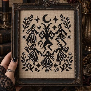May include: A framed cross-stitch embroidery featuring a black thread design on a beige fabric background. The pattern depicts a central horned figure surrounded by four dancing individuals, accented with stars, a crescent moon, and floral motifs.