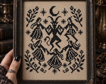 May include: A framed cross-stitch embroidery featuring a black thread design on a beige fabric background. The pattern depicts a central horned figure surrounded by four dancing individuals, accented with stars, a crescent moon, and floral motifs.