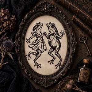 May include: An oval-framed cross-stitch embroidery featuring two frogs dancing. One frog wears a dress and the other wears a tuxedo with a bow tie. The design is rendered in black thread on a light beige fabric, encased in an ornate dark brown frame.