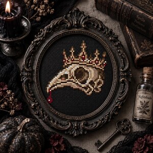 May include: A cross-stitch embroidery featuring a beige bird skull wearing a crown with red accents, set against a black fabric background. The piece is encased in an ornate, dark brown circular frame with intricate carved detailing along the edges.