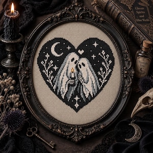 May include: A cross-stitch embroidery in an oval frame featuring two ghosts under a sheet holding a candle inside a black heart shape. The design includes a crescent moon, small stars, and floral sprigs on a beige fabric background.
