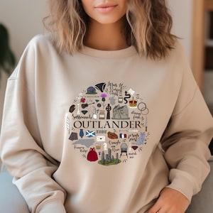 Può includere: Felpa beige con un disegno circolare con la parola "OUTLANDER" e varie illustrazioni. Il design include testo e immagini come una croce, un vestito e la frase "Jesus H. Roosevelt Christ."
