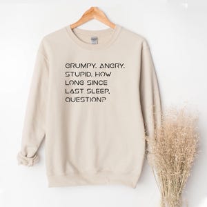 May include: A beige sweatshirt with the text "GRUMPY. ANGRY. STUPID. HOW LONG SINCE LAST SLEEP, QUESTION?" in black. The sweatshirt is displayed on a wooden hanger against a white background.