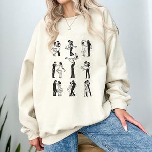 May include: A beige sweatshirt featuring a repeating pattern of black and white dancing couples. The graphic depicts various dance poses, including lifts and embraces. The sweatshirt has a crew neck and long sleeves.