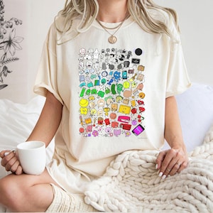 May include: Cream-colored t-shirt featuring a colorful, whimsical illustration of various cartoon characters and objects. The design includes a diverse array of smiling faces, food items, and tech gadgets. The shirt is worn by a person holding a white mug.