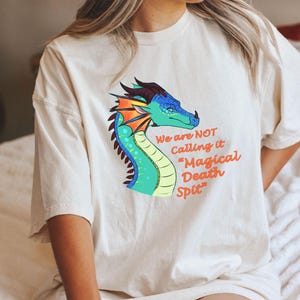 May include: A cream-coloured t-shirt with a dragon illustration in blue, green, orange, and red, and the text "We are NOT Calling it Magical Death Spit". The shirt is a relaxed fit, suitable for casual wear.