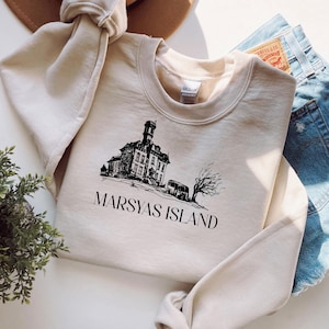 Könnte beinhalten: Beiges Sweatshirt mit schwarzer Grafik eines Gebäudes, eines Busses und eines Baumes, darunter der Text "MARSYAS ISLAND". Das Sweatshirt ist gefaltet, ein Teil einer Jeansshorts ist sichtbar.
