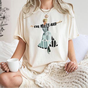 May include: Cream-colored t-shirt featuring the text "THE HILLS ARE ALIVE WITH THE SOUND OF MUSIC" in black and teal. The graphic depicts a woman with outstretched arms. The shirt is a relaxed fit.