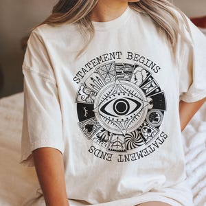 May include: Cream-colored t-shirt featuring a circular black graphic with an eye at the center and the words "STATEMENT BEGINS" and "STATEMENT ENDS". The graphic includes various symbols and illustrations.