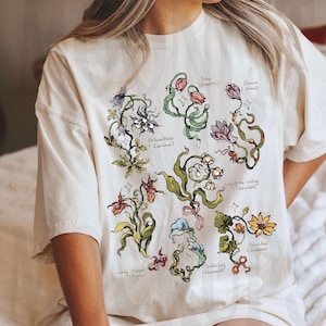 Pode incluir: Uma t-shirt de manga curta cor de creme com uma ilustração botânica de várias flores, incluindo tulipa, açafrão, aquilégia, lírio-do-vale, sapatinho-de-senhora, copo-de-leite e celidónia, cada uma rotulada com o nome e um nome entre parênteses.