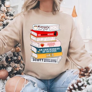 May include: Beige sweatshirt featuring a stack of books with various titles, including "Karen's Cafe," "Red Bedroom Records," and "An Unkindness of Ravens." The design is a colorful graphic, perfect for book lovers and those who enjoy unique apparel.