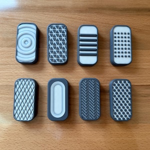 May include: An assortment of eight rectangular, patterned objects with a dark gray border and white or gray designs. The patterns include concentric circles, triangles, horizontal lines, dots, and other geometric shapes. The objects are arranged on a wooden surface.